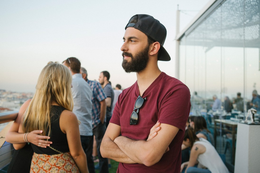 Man in maroon shirt and cap standing on a rooftop terrace with other guests and cityscape views behind him.