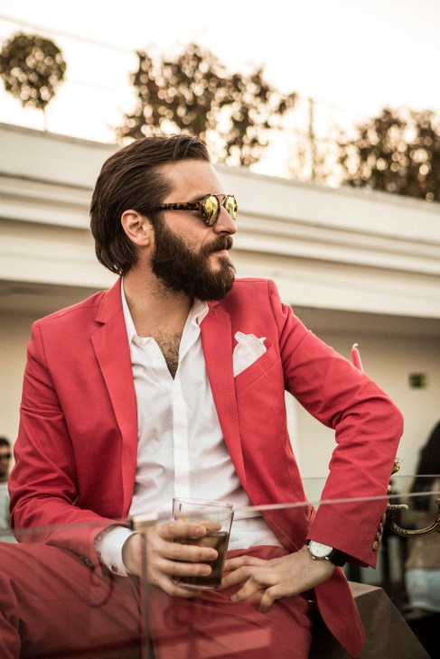 Man in red blazer and white shirt wearing mirrored sunglasses holds a drink at an outdoor venue.