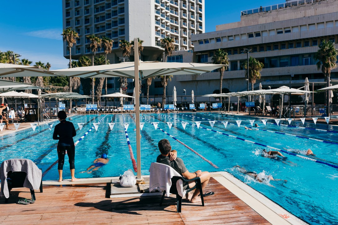 Olympic-size swimming pool with lane dividers and swimmers, surrounded by modern buildings and palm trees in Tel Aviv