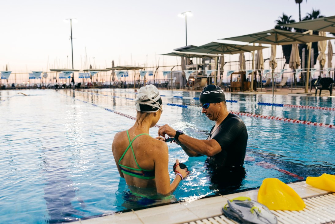 Two swimmers in a pool adjusting swimming equipment, one wearing a green swimsuit and swim cap