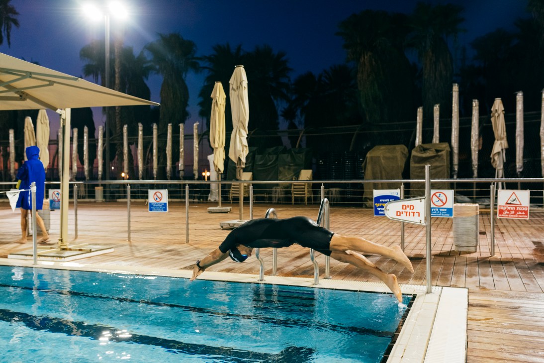 Person diving into outdoor swimming pool at night with wooden deck and umbrellas in background