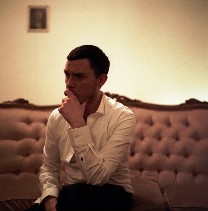 Man in white shirt sitting on beige couch with hand to face in contemplative pose