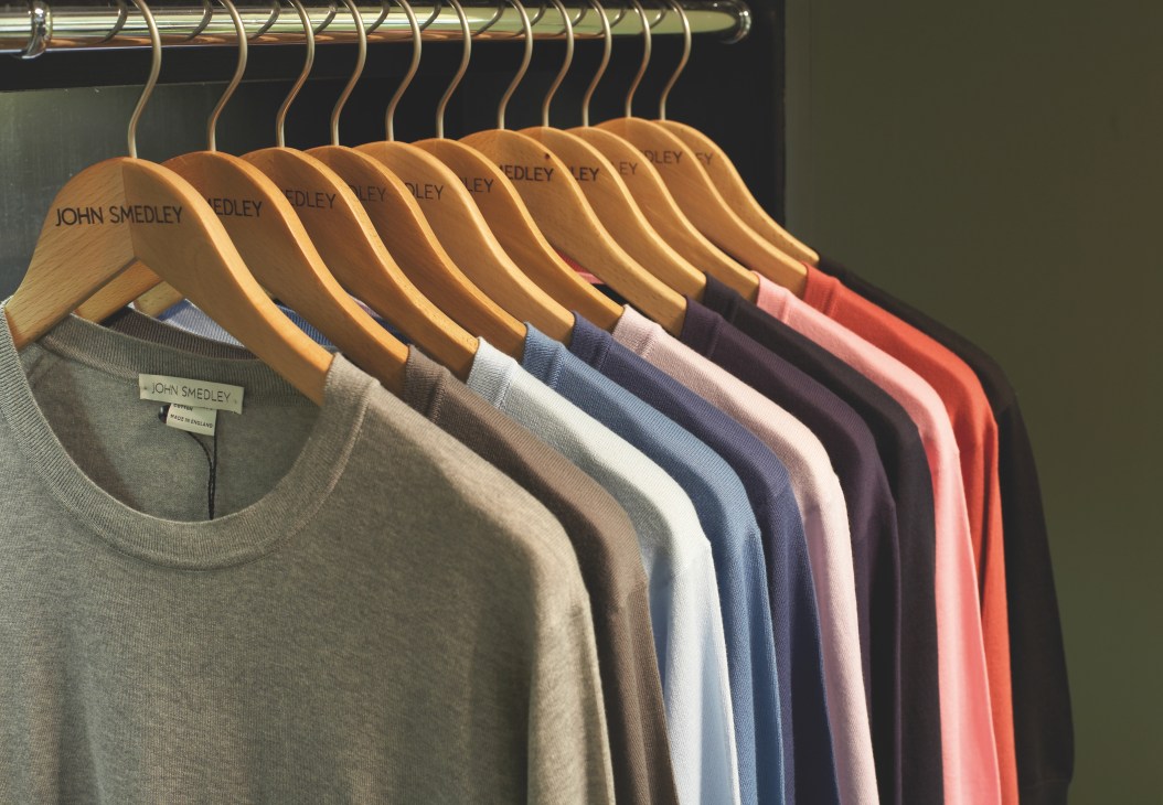 Row of John Smedley knitwear in various colors hanging on wooden hangers with brass rail.
