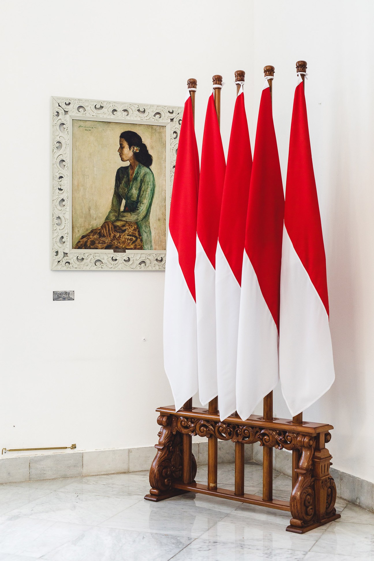 Indonesian red-and-white flags displayed on an ornate wooden stand next to a framed portrait painting on a white palace wall.