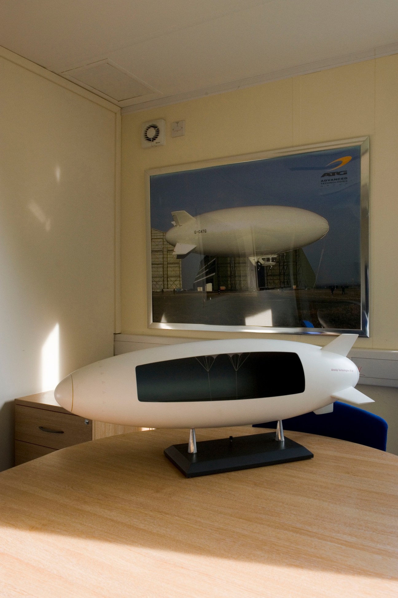 Scale model of a hybrid airship with white fuselage and black gondola displayed on a wooden desk in an office.