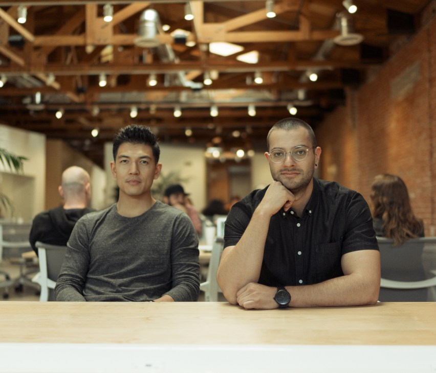 Two men sitting at a table in a modern office space with exposed wooden beams and industrial lighting.