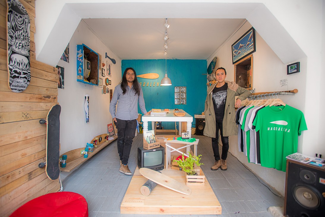 Two people stand in a narrow retail shop with wooden walls, displaying skateboards, clothing, and surf/skate merchandise.