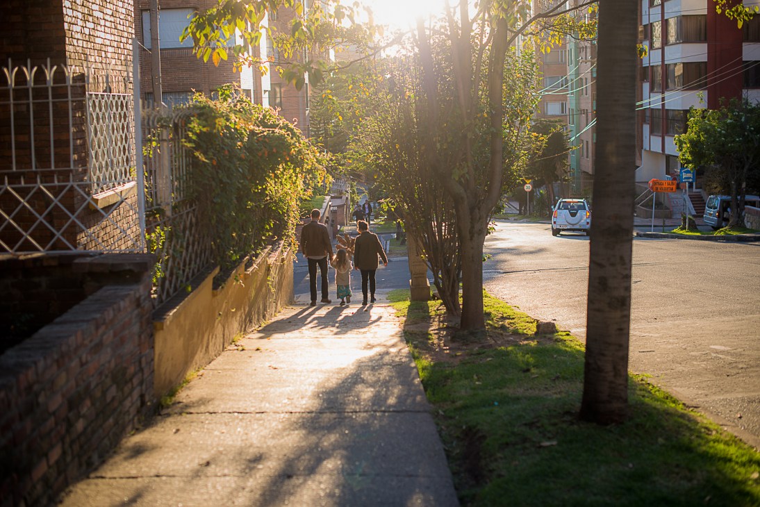 Tree-lined street in Chapinero Alto with residents walking, brick buildings, and dappled sunlight filtering through foliage.