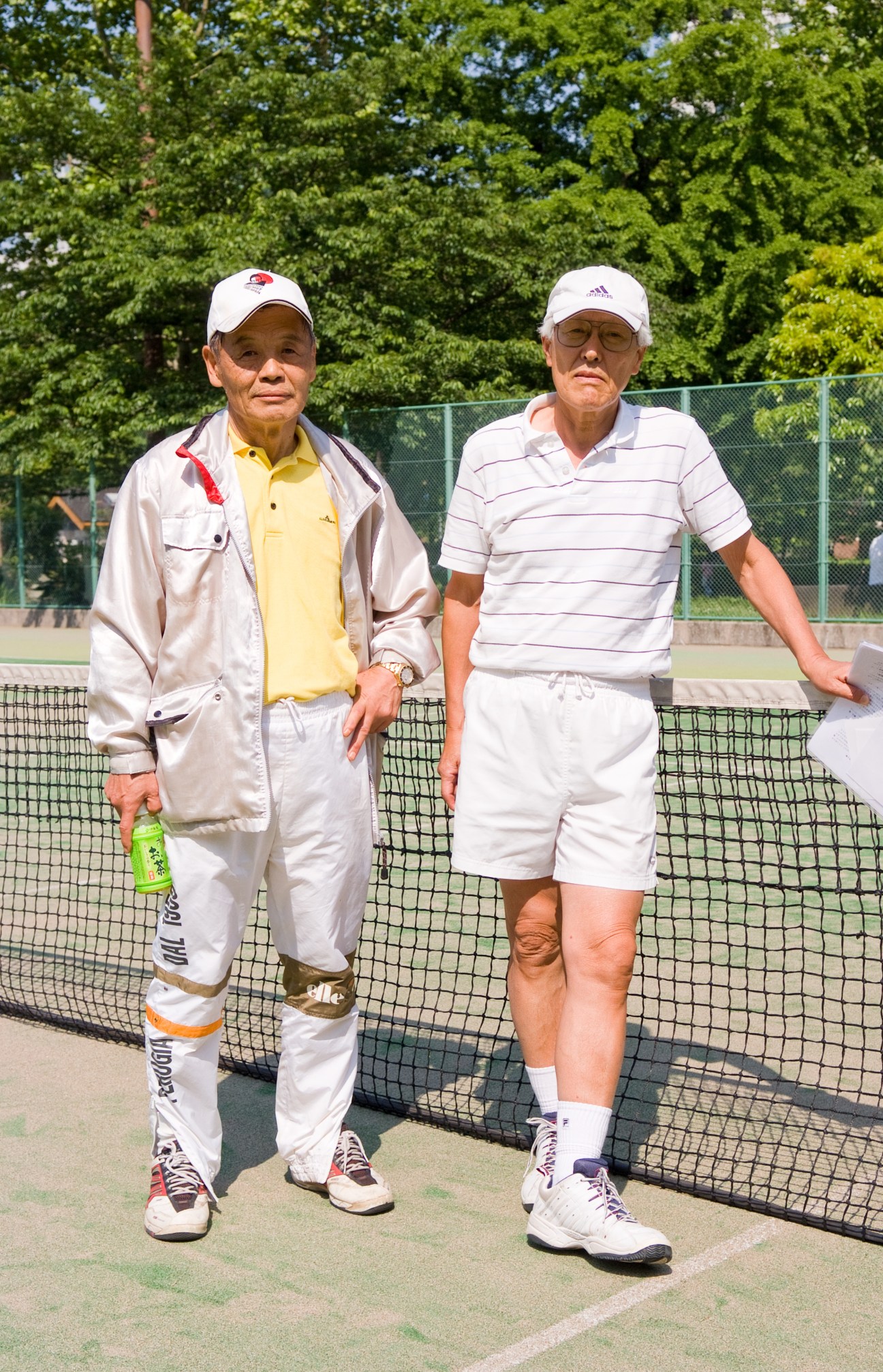 Two older Japanese men in tennis attire standing by a net on a tennis court.