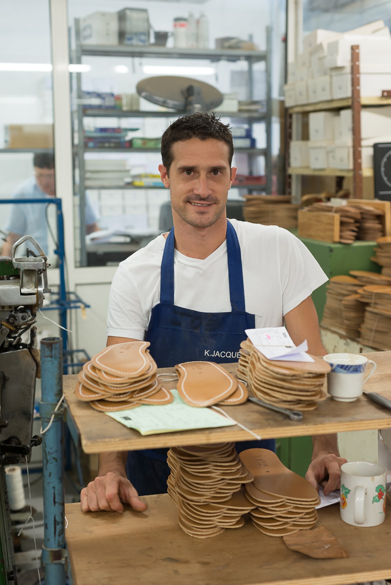 K-Jacques worker in blue apron counting stacks of leather sandal soles at workbench.