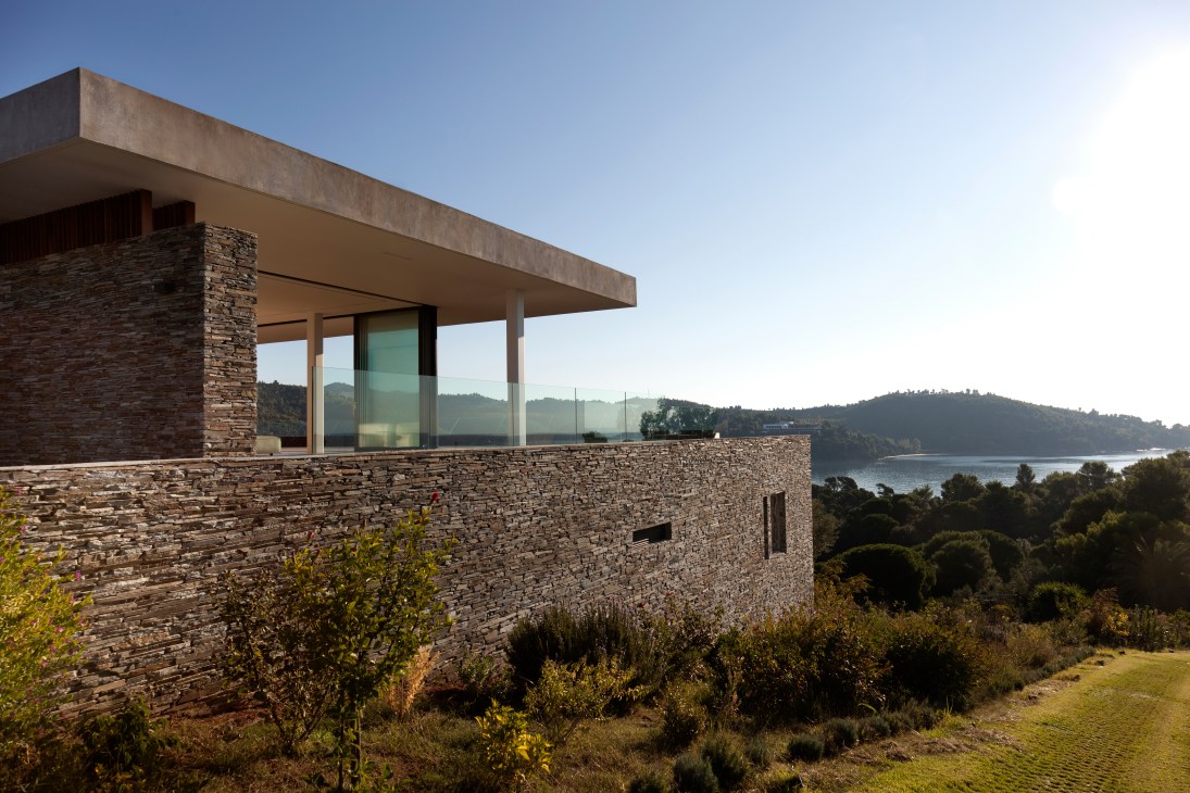 Modern house with stone walls and cantilevered concrete roof overlooking a lake and forested hills.
