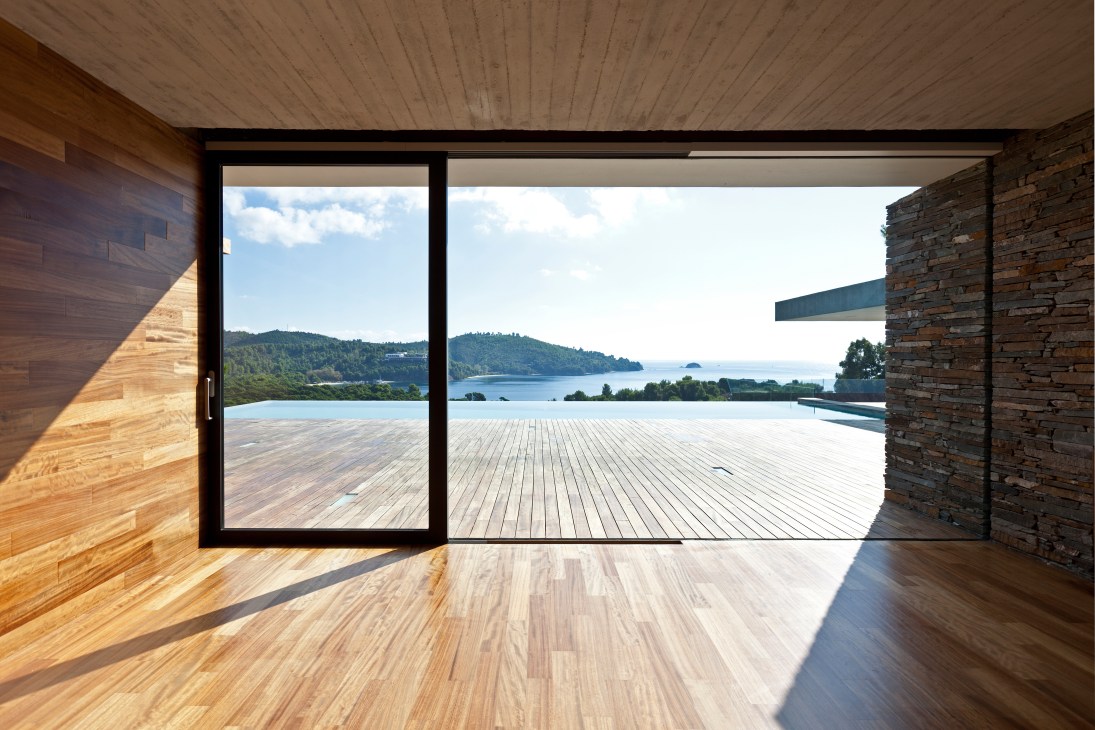 Interior view of Plane House master bedroom with wooden floors, stone walls, and expansive windows overlooking a coastal landscape.