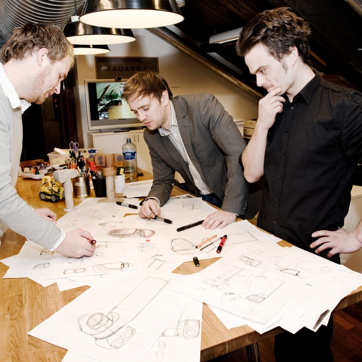 Three men review design sketches at a wooden table in a modern office space.