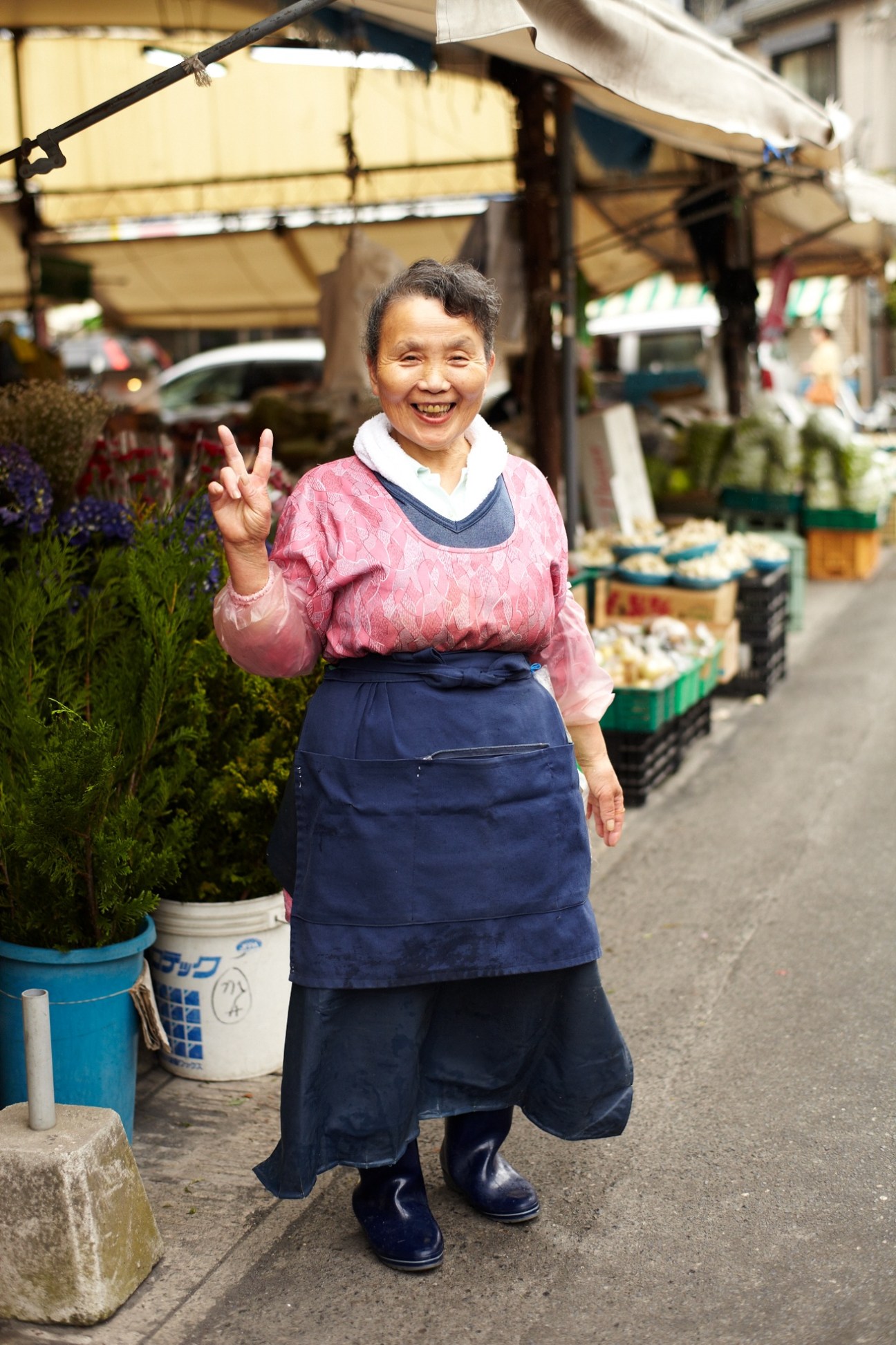 Smiling woman vendor at an outdoor market stall wearing pink and blue clothing, making a peace sign gesture.