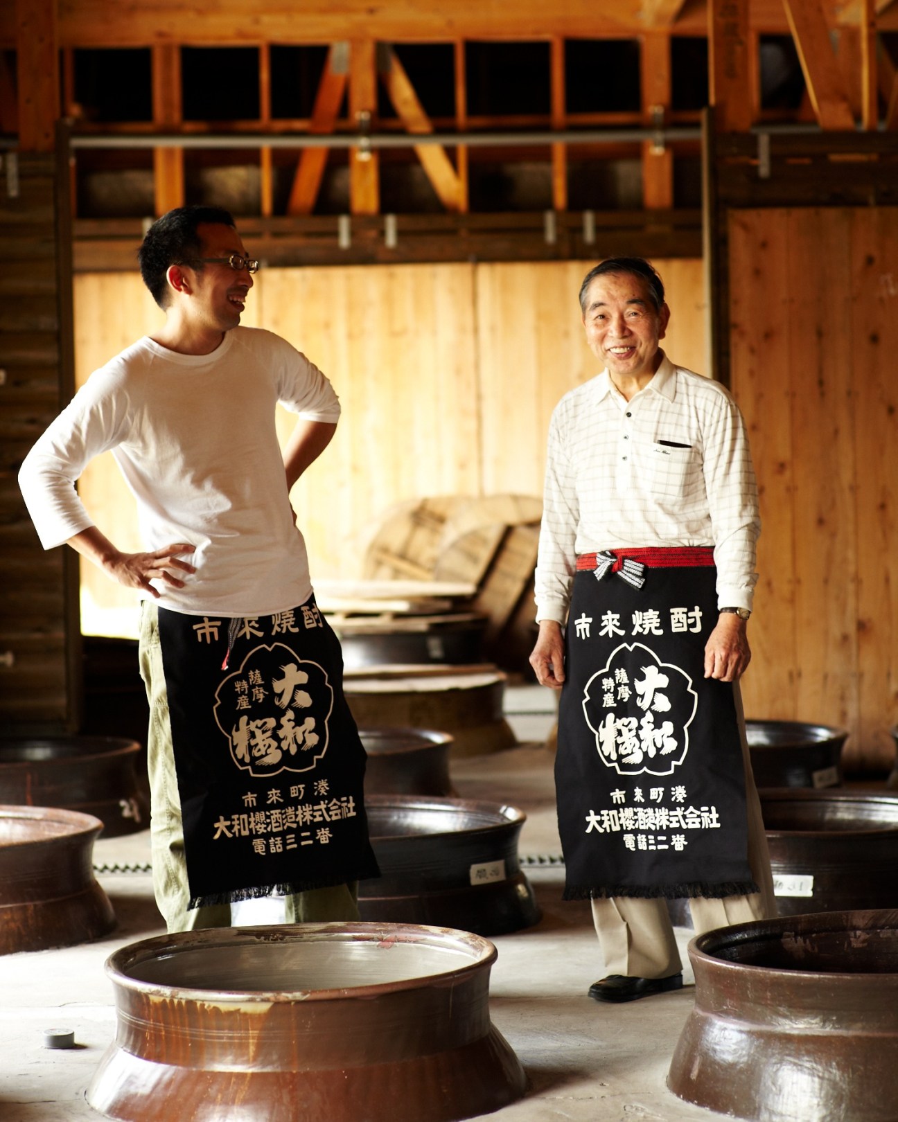 Kazunori Wakamatsu and his son Tekkan Wakamatsu stand in a sake brewery wearing traditional aprons, with fermentation vessels visible.