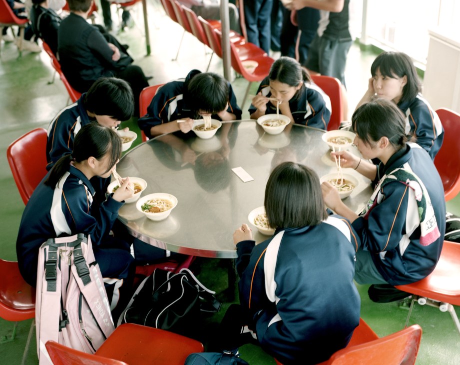Schoolchildren in uniforms eating udon noodles at a round table with red chairs on a ferry.