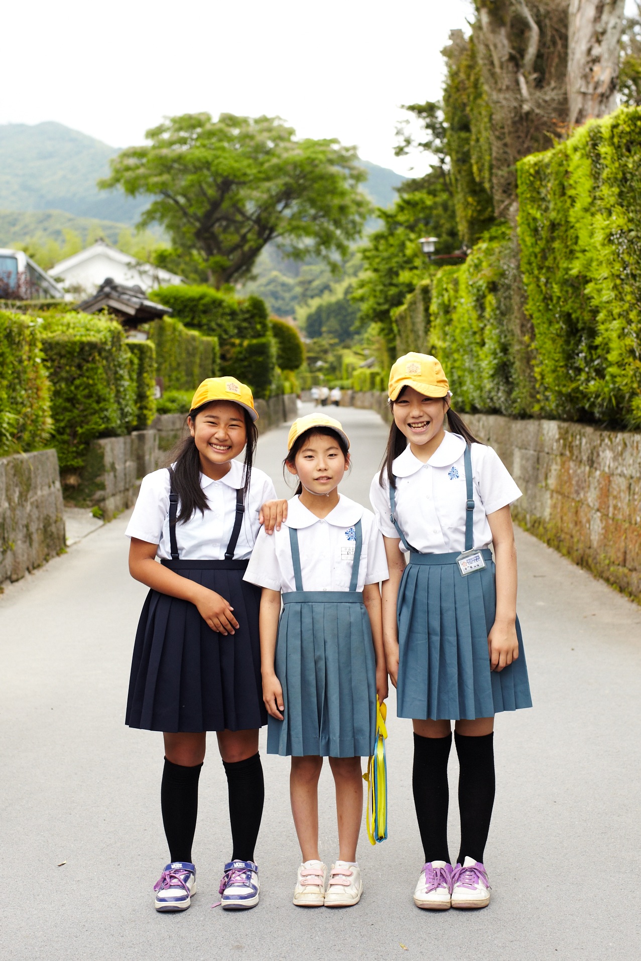 Three Japanese schoolgirls in uniforms and yellow caps standing on a tree-lined street in Chiran.