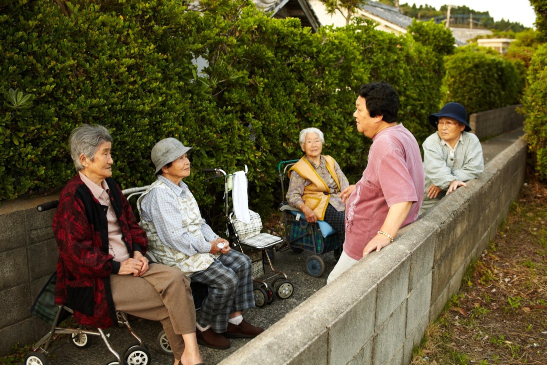 Five elderly Japanese people chatting outdoors by a concrete wall with green hedges behind them.