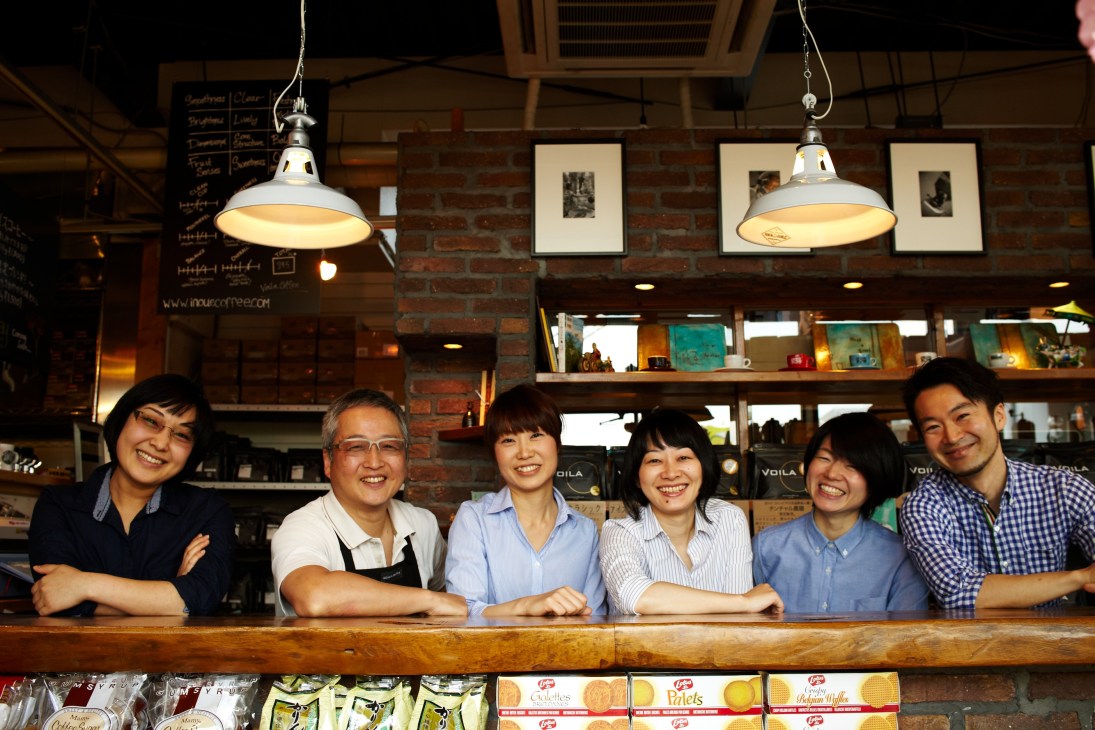 Voila café owner Tatsuya Inoue and staff smiling behind counter under pendant lights.