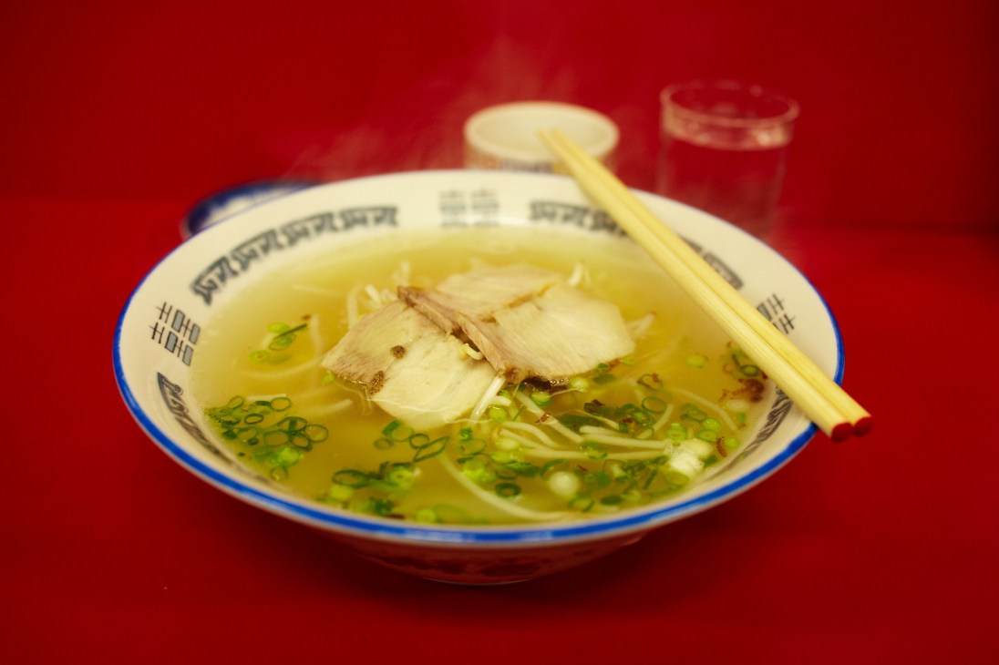 Bowl of ramen with noodles, broth, sliced pork and green onions with chopsticks against red background.