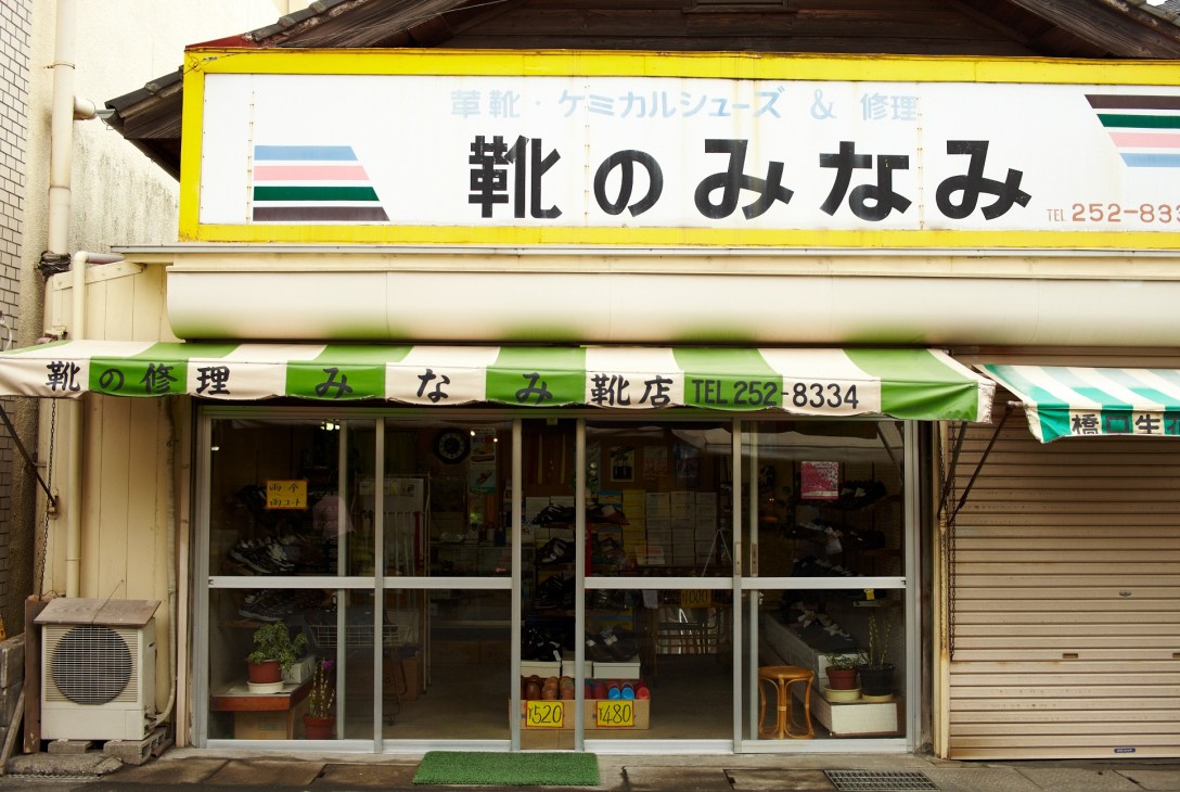 A shoe repair shop storefront with yellow and green signage displaying Japanese characters near Kagoshima Central station.