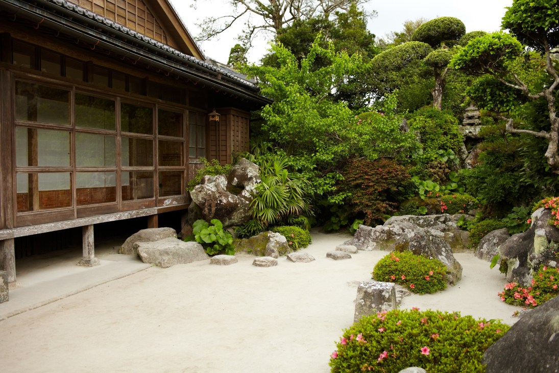 Traditional samurai house with wooden facade and shoji windows overlooking a Japanese garden with rocks, moss, and flowering plants.