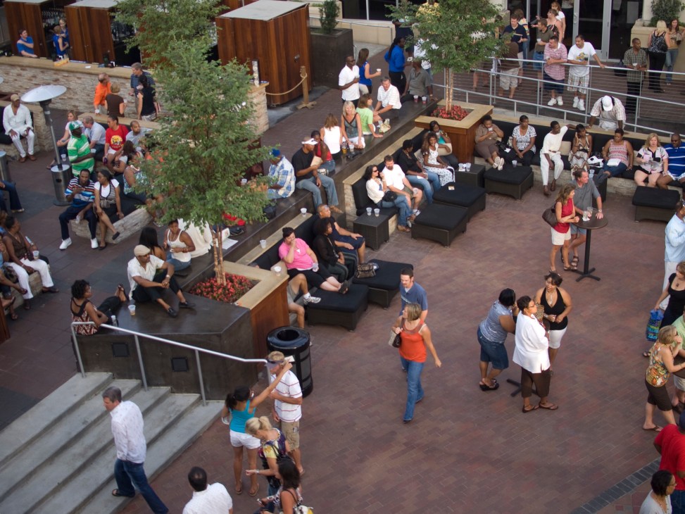 Aerial view of diverse crowds gathered in an outdoor urban plaza with seating and trees.