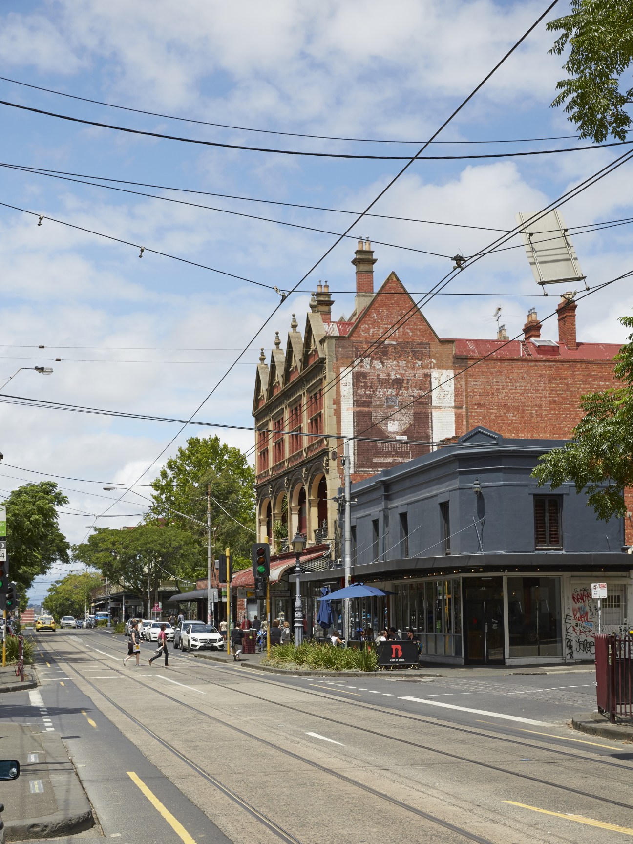 A wide view of Gertrude Street in Melbourne's Fitzroy, showing Victorian terraces, power lines, tram tracks, and pedestrians shopping on a sunny day.