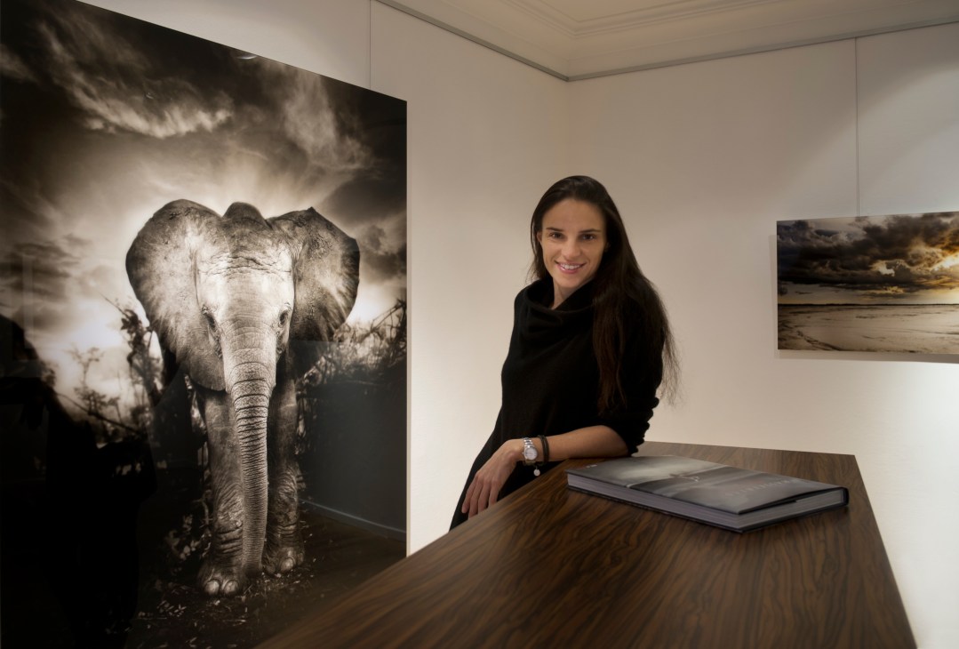 Kathrin Köhler, founder of Immagis gallery, poses at a wooden desk next to a large black-and-white photograph of an elephant.