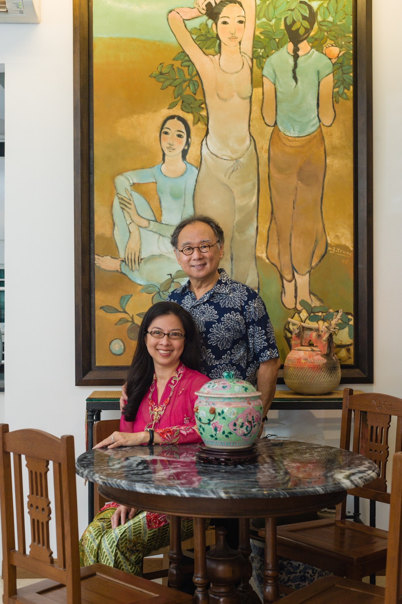 Colin and Linda Chee sit at a round marble table with wooden chairs in front of a large figurative painting.