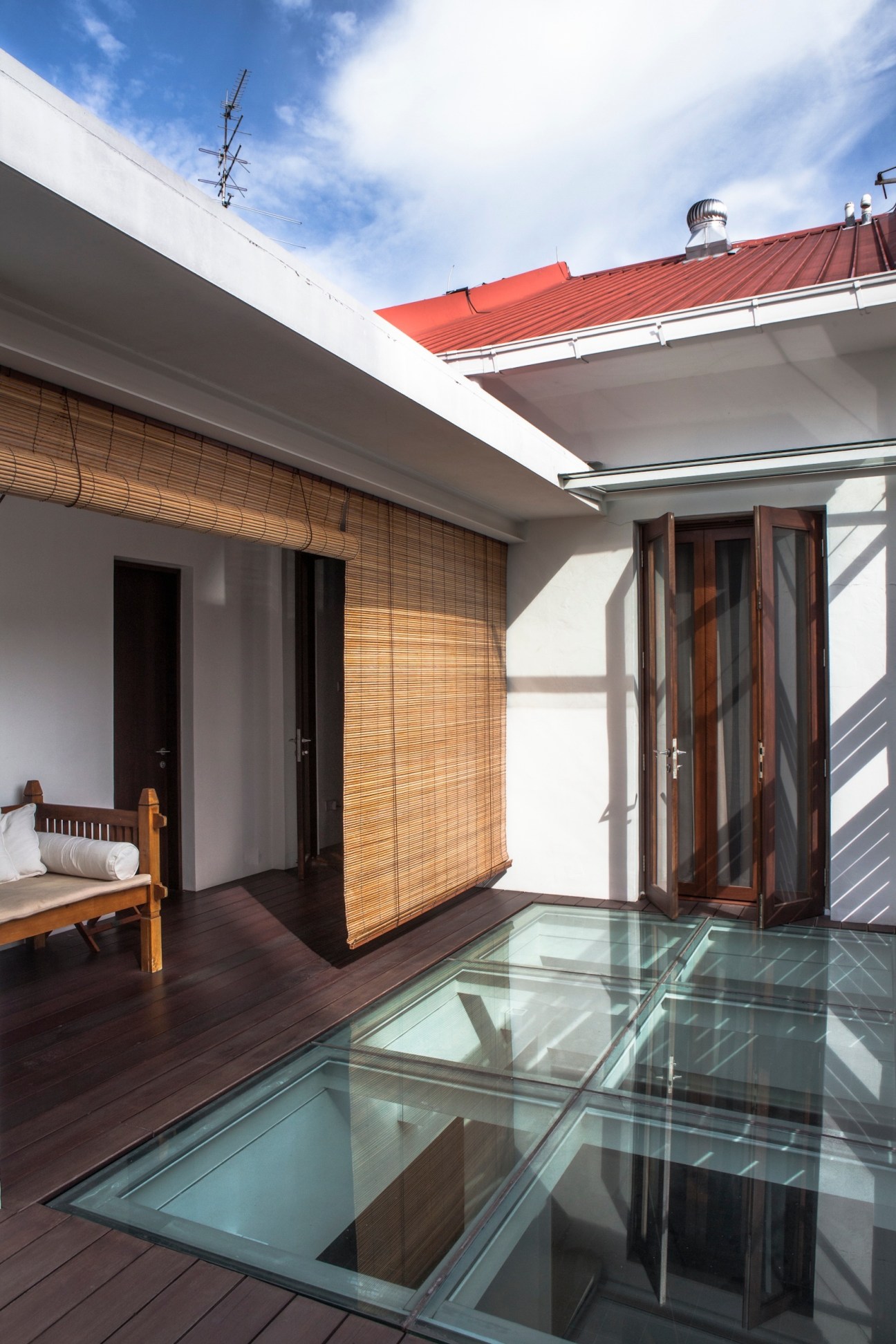 Modern rooftop patio with wooden deck, glass skylight, bamboo screens, and red-roofed building against blue sky.
