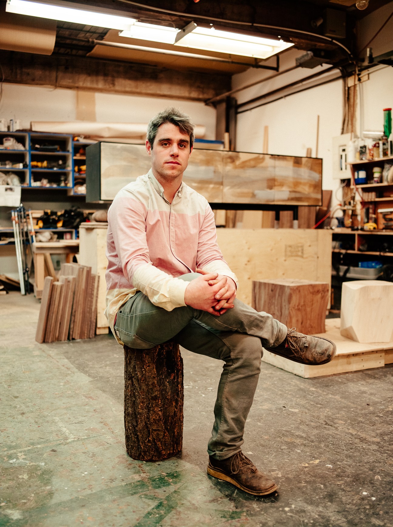 Wood-furniture designer Jeff Martin sitting on a log in his workshop surrounded by wooden materials and tools.