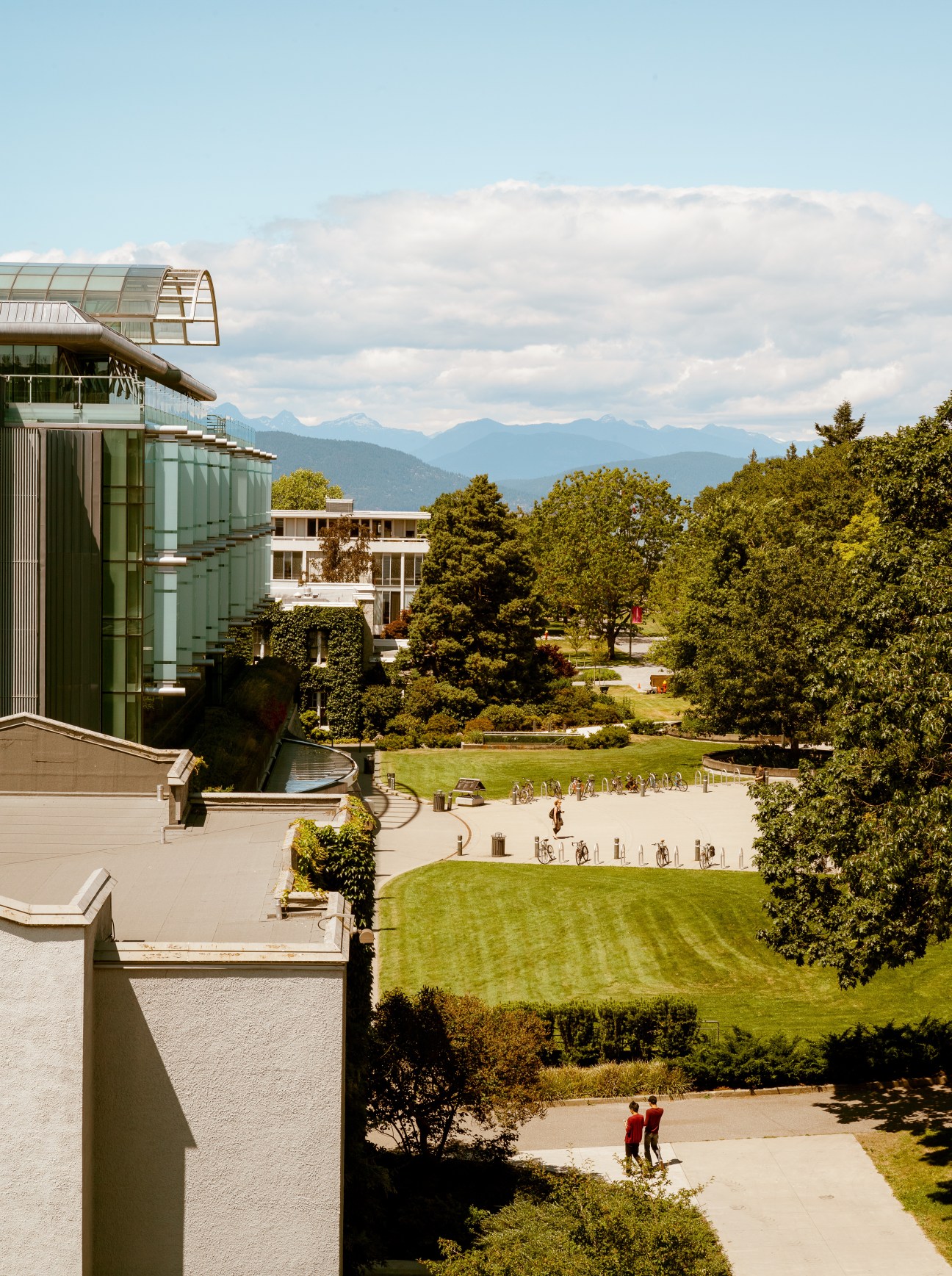UBC Sauder School campus overlooking manicured lawns and modern buildings with North Shore mountains in distance.