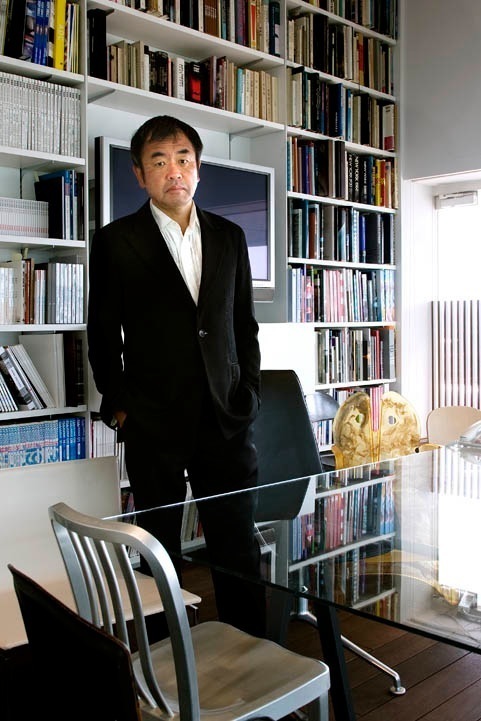 Architect Kengo Kuma stands in his office beside a desk with bookshelves behind him.