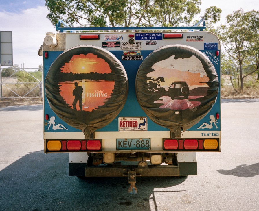 Rear view of a blue and cream campervan with large spare tire covers decorated with outback scenes and 'Gone Fishing' text.