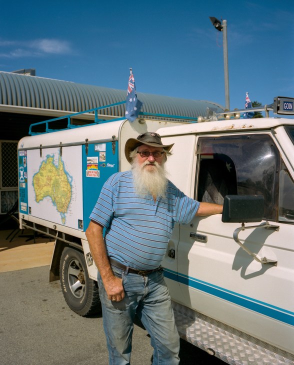 Retired man with white beard standing beside campervan decorated with Australian map and flag.