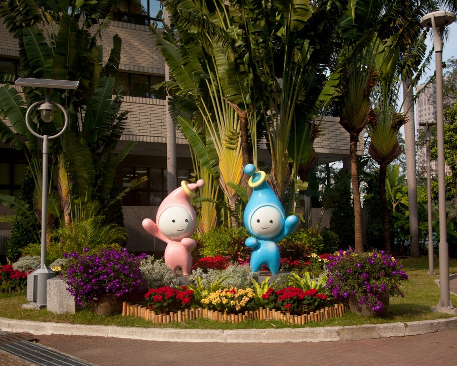 Two colorful mascot characters stand among tropical plants and flowering shrubs outside a modern building.