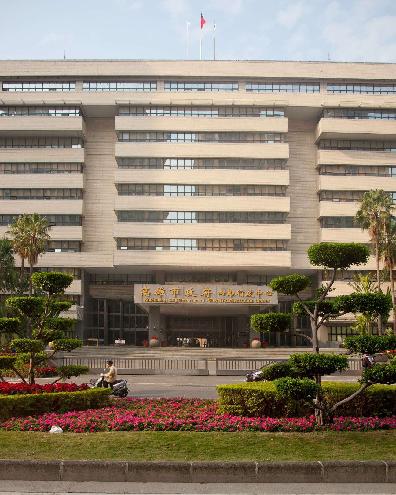 Kaohsiung city hall, a modernist multi-story government building with horizontal bands of windows and Chinese characters on the facade, surrounded by landscaped gardens with red flowers.