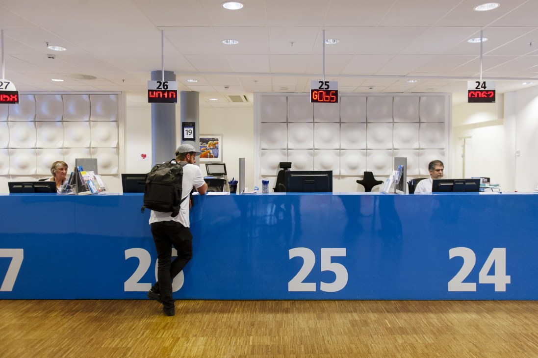 Man at service counter in modern white and blue office with staff at numbered stations.