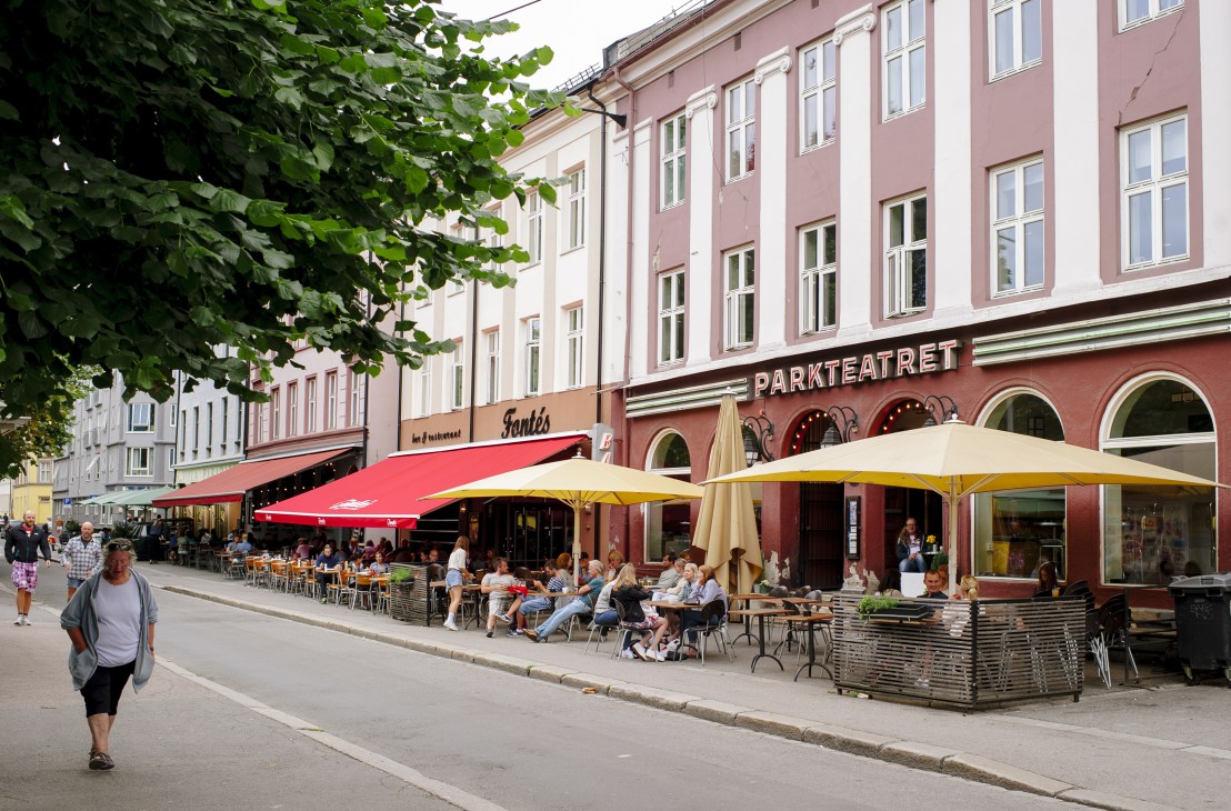 Oslo street with outdoor café seating, pastel-colored buildings, and tree-lined pedestrian area.