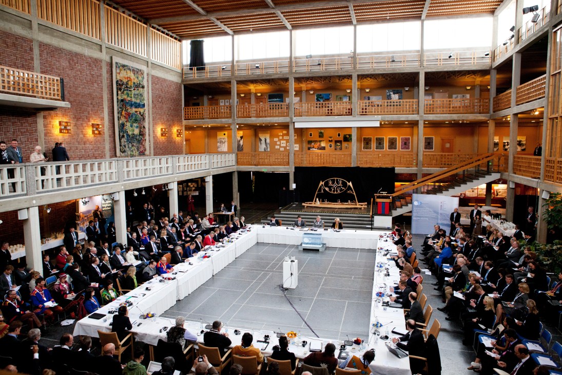 Interior view of Kiruna town hall with Arctic Council delegates seated at long white tables in a modern wooden and brick assembly hall.
