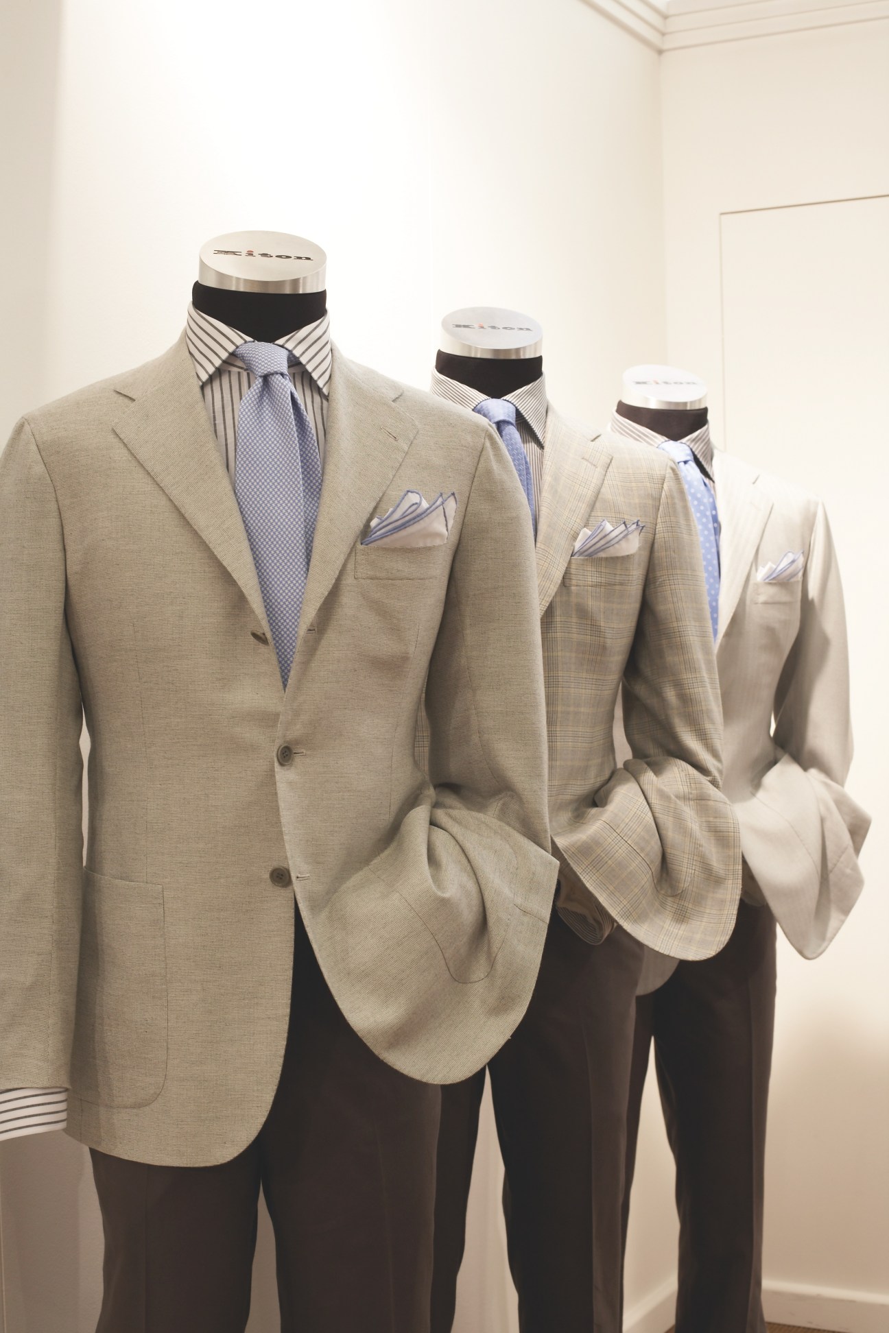 Three tailored suits by Kiton displayed on mannequins, featuring beige blazers with striped shirts and patterned ties.