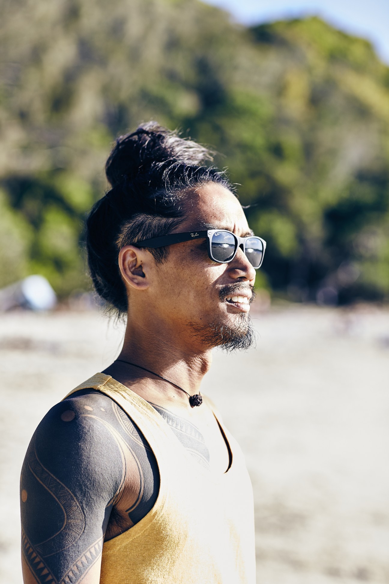 Man wearing sunglasses and casual beachwear stands on a sunny beach with green cliffs in the background.