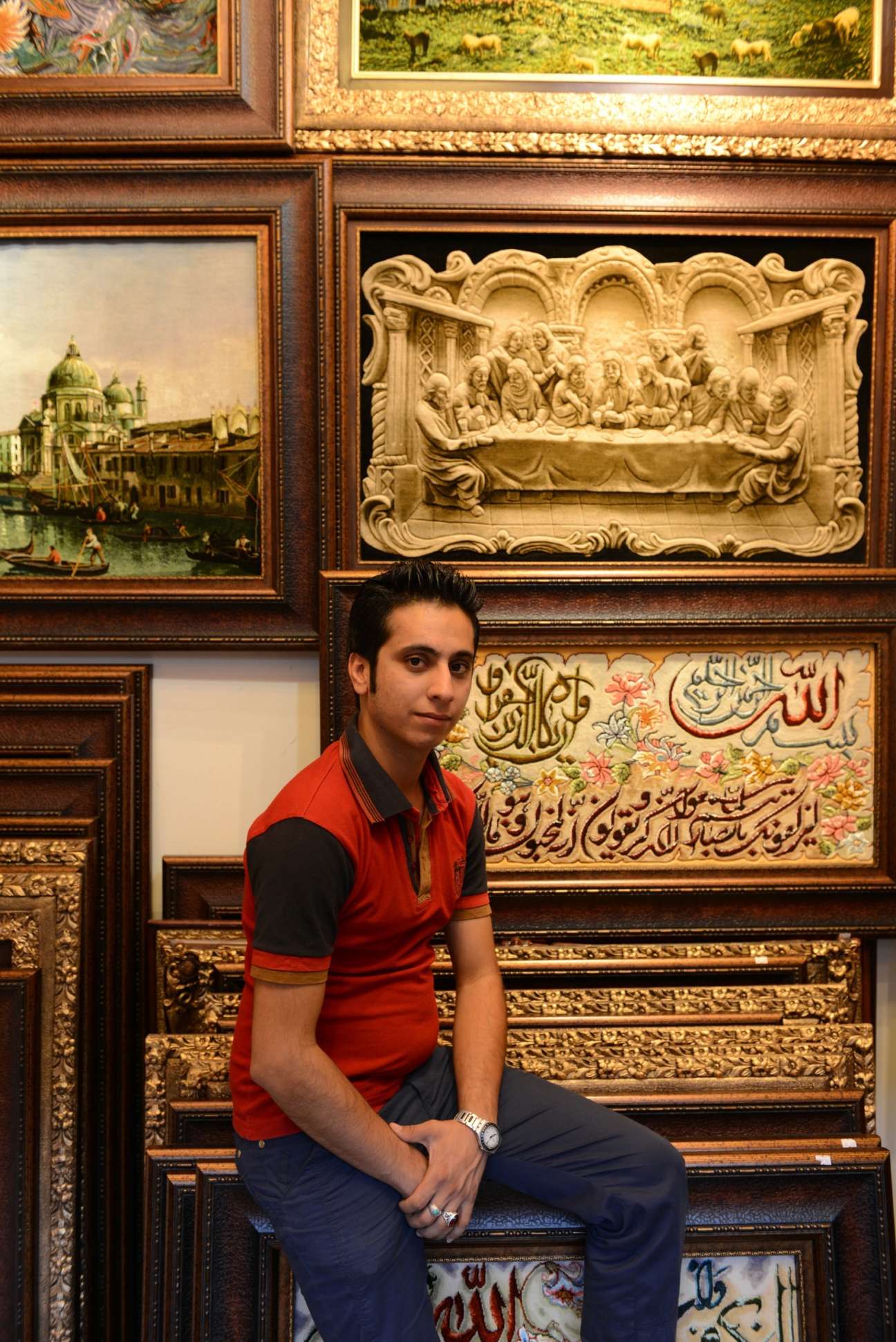 Rasoul, a carpet bazaar vendor, sits against ornate wooden frames displaying religious artwork and decorative panels.