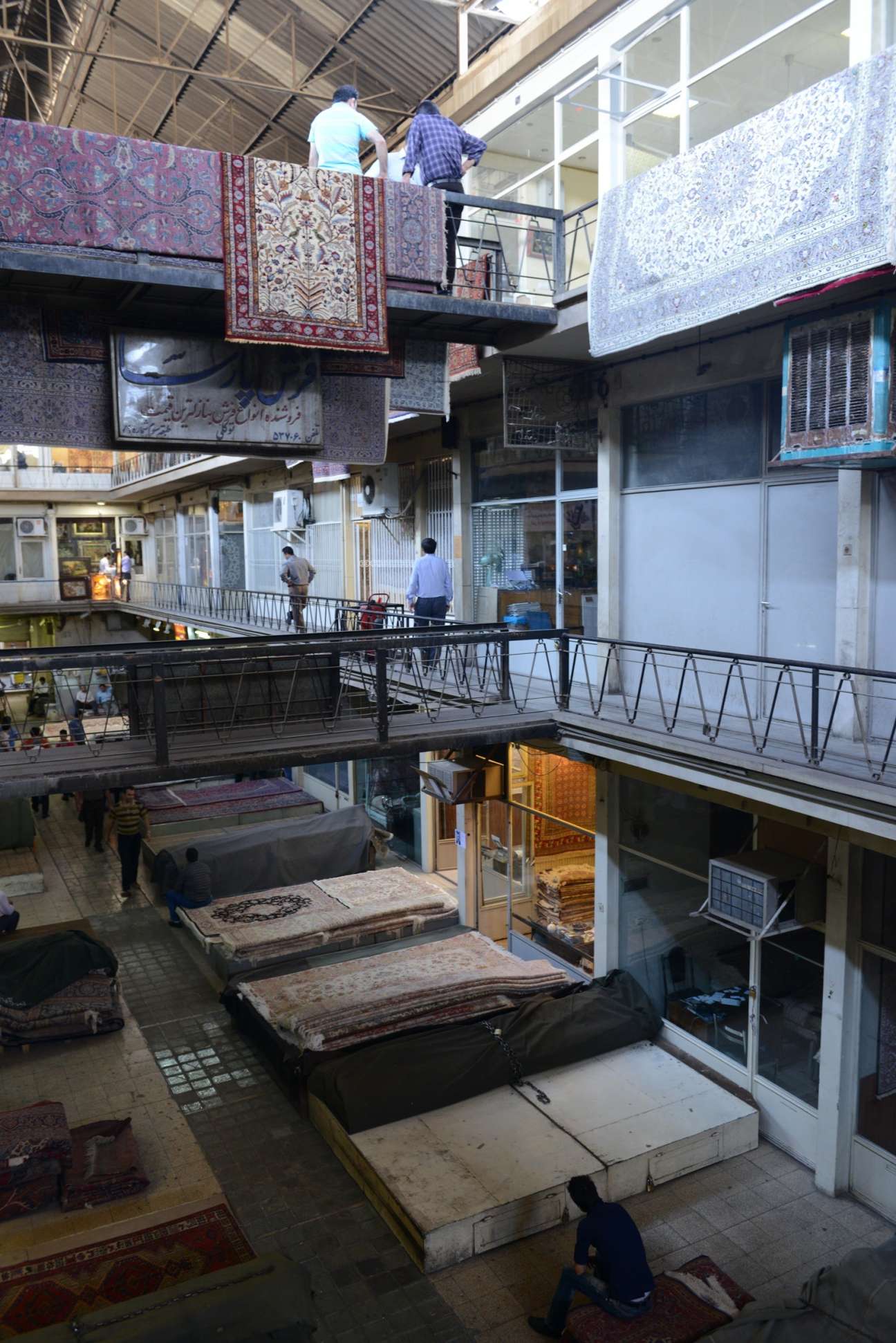 Multi-level carpet bazaar interior with rolled carpets displayed on platforms and vendors on overhead walkways.