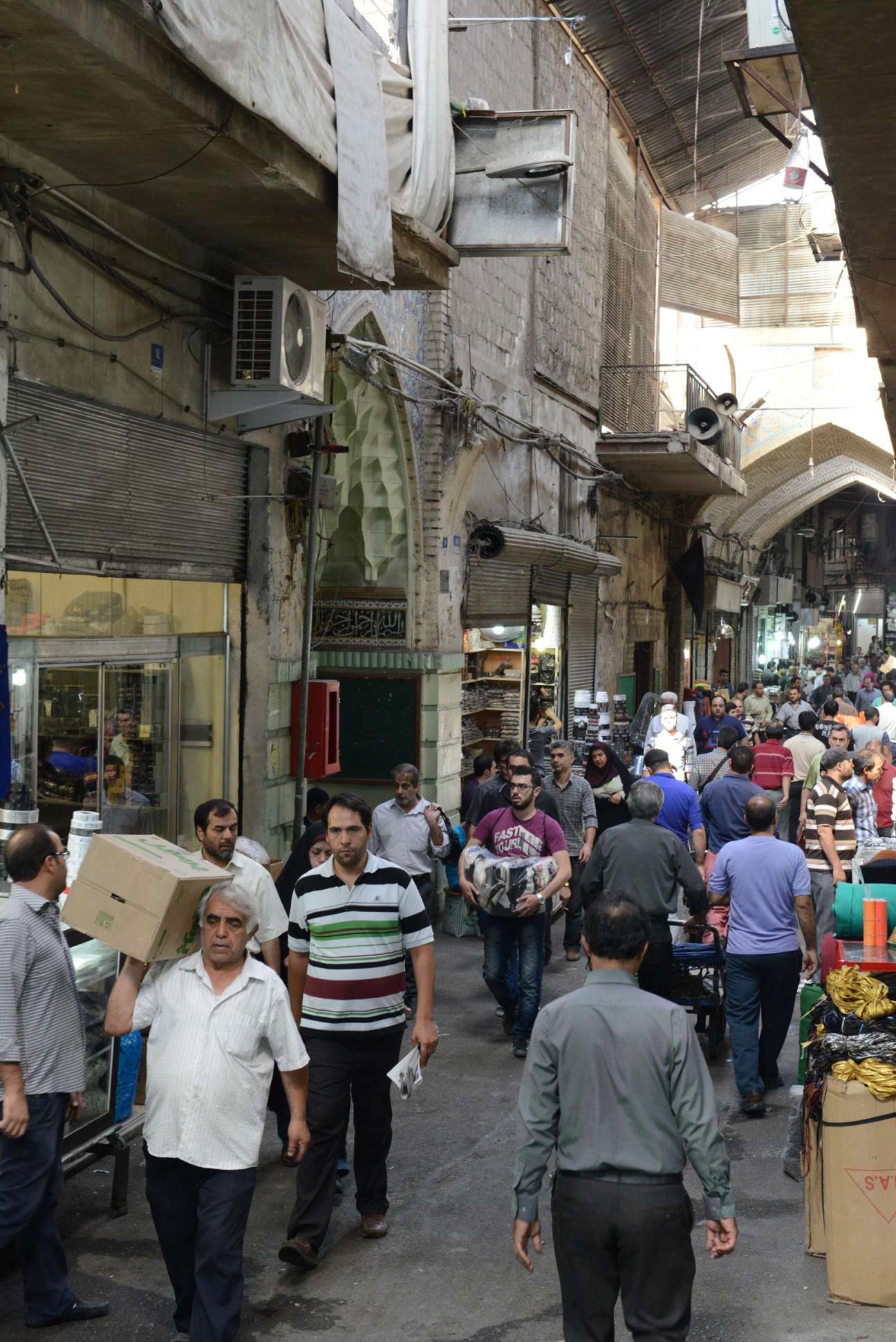 Crowded street scene inside Tehran's Grand Bazaar with shoppers and vendors among market stalls and buildings.
