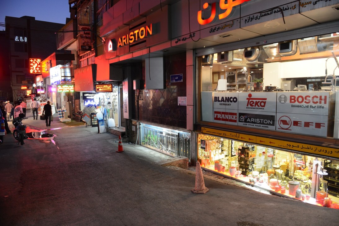 Night street scene in North Tehran with illuminated shop fronts selling kitchen and bathroom suites, displaying brand names like Ariston and Bosch.