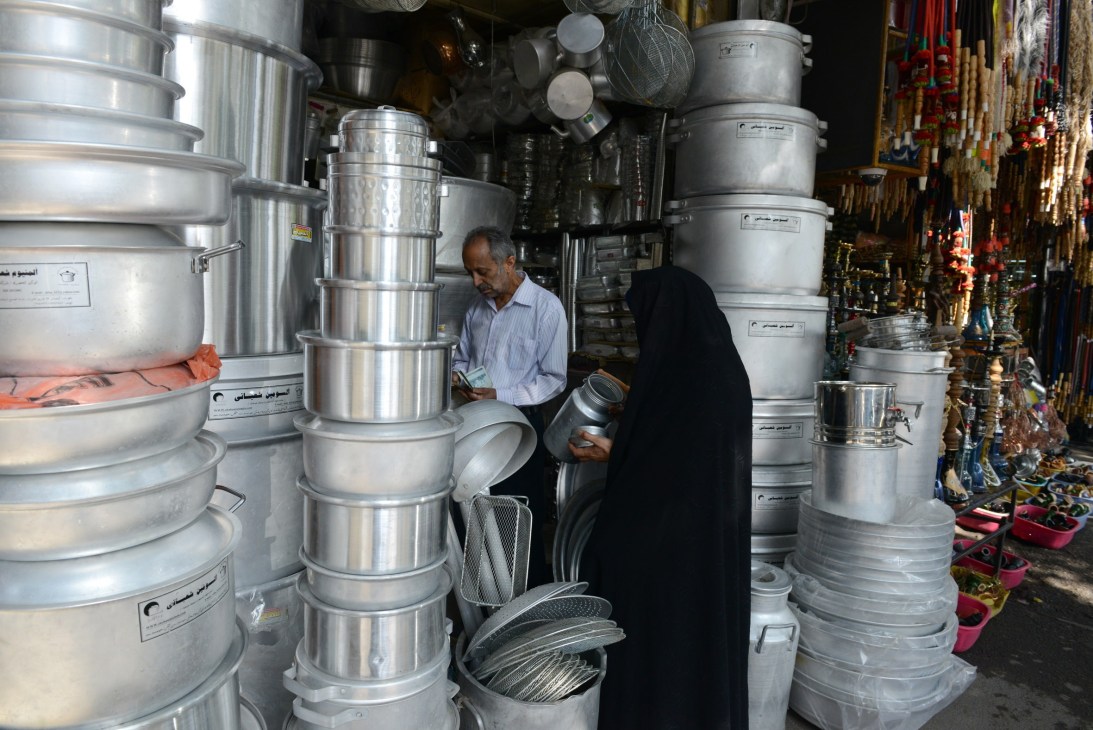 A woman in a black chador browses aluminium cookware and kitchenware stacked high in a bazaar shop.