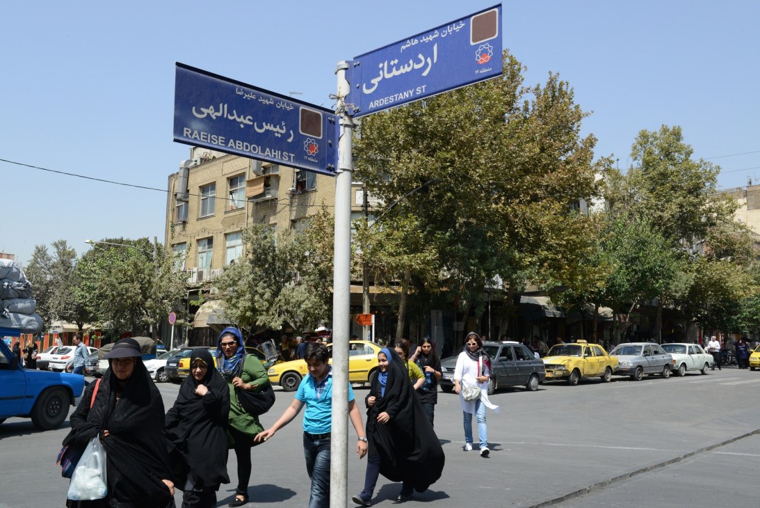 A boy in blue walks among women wearing black chadors on a Tehran street corner with bilingual street signs.