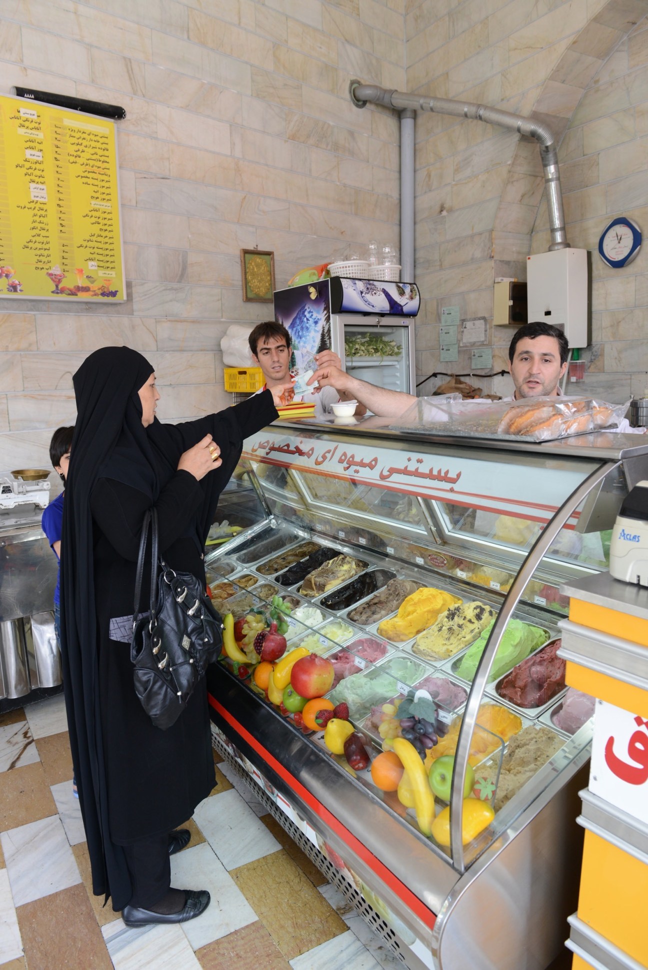 Woman in black chador ordering ice cream from male vendor at counter in Tehran shop